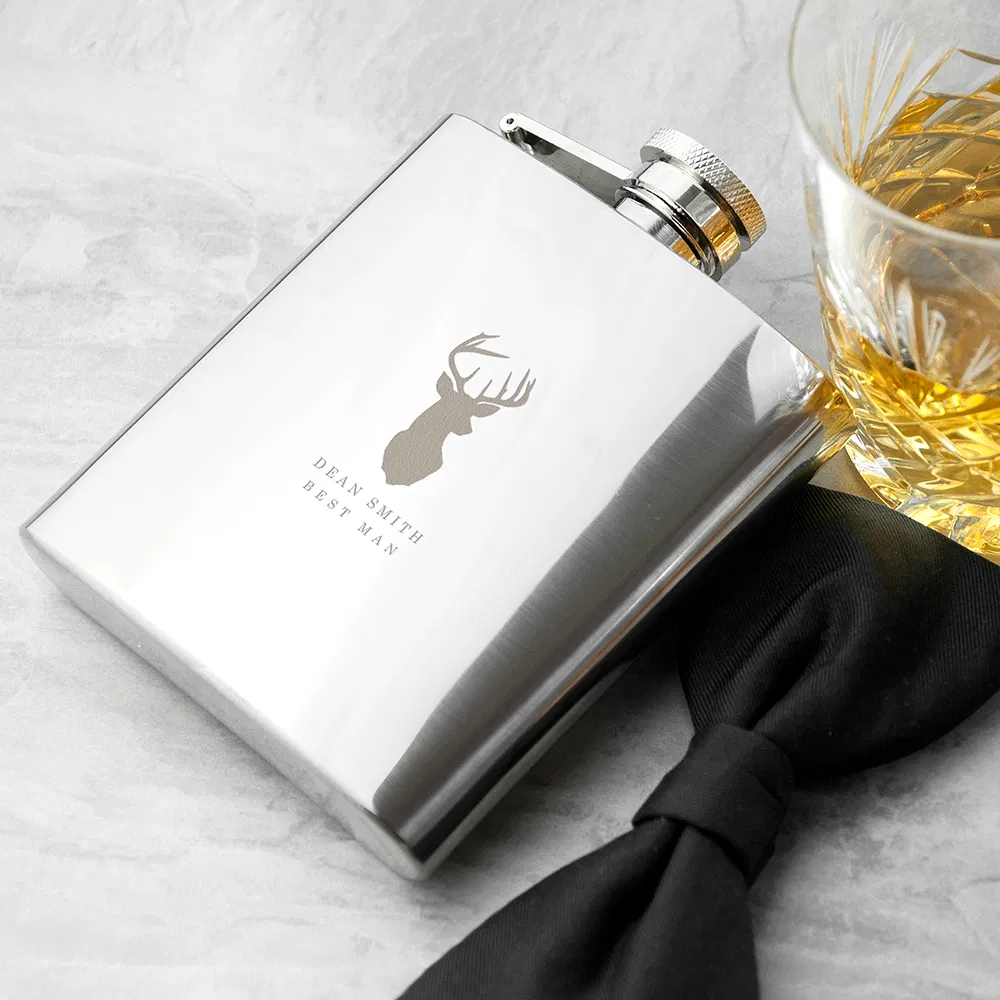 Polished stainless steel 6oz hip flask featuring an engraved stag head motif and custom text, styled on a grey surface next to a glass of whisky and a black bow tie.
