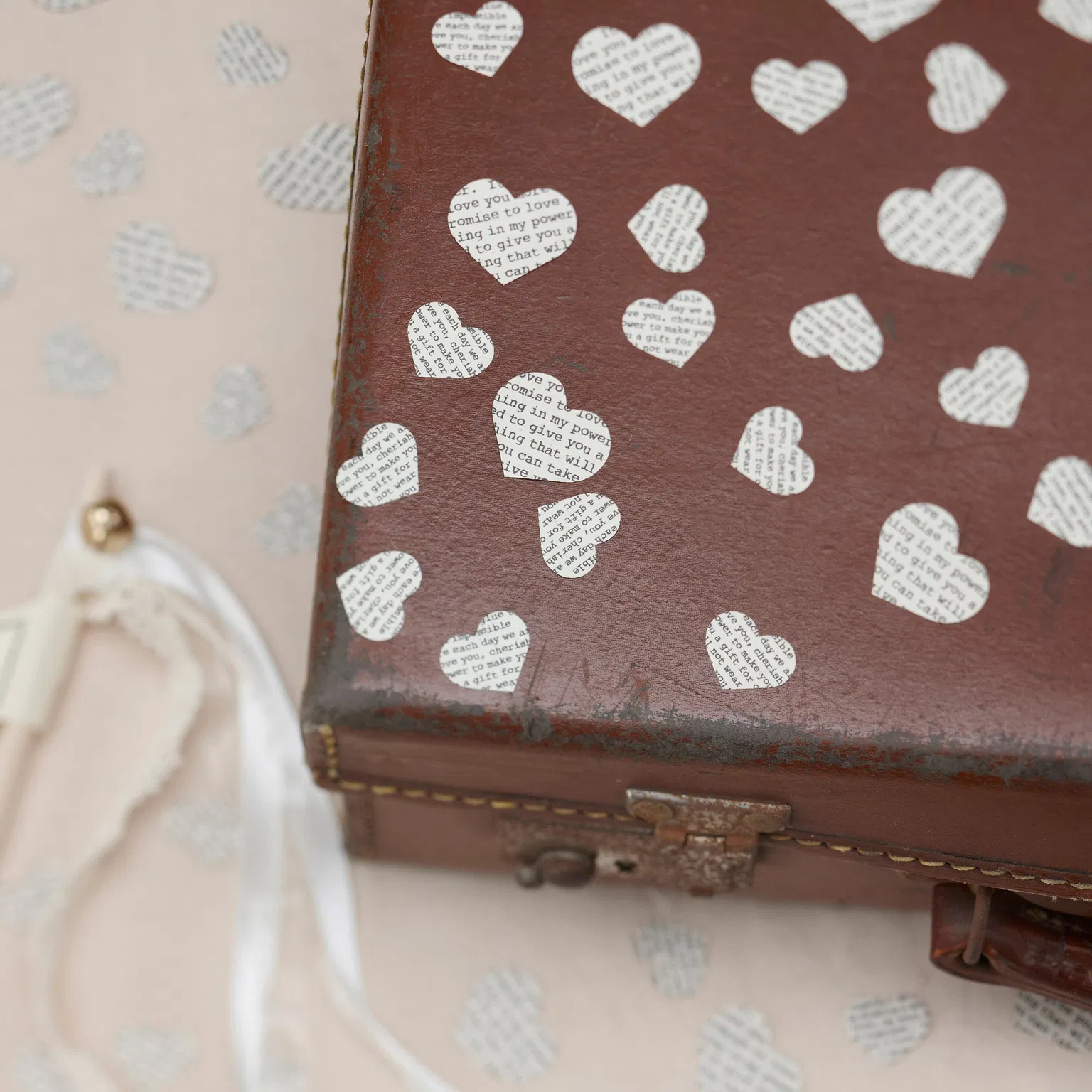 Vintage heart-shaped confetti with printed love poem text scattered over a rustic brown leather suitcase with white ribbons.