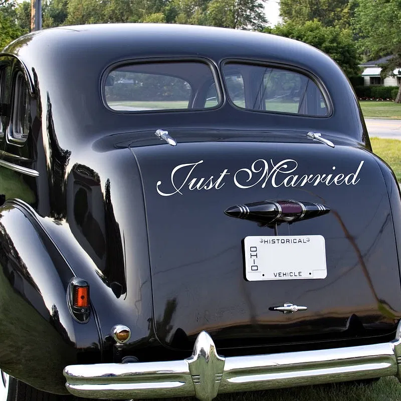Classic black vintage car with 'Just Married' engraved in elegant white script vinyl on the rear trunk, parked on a sunlit street.