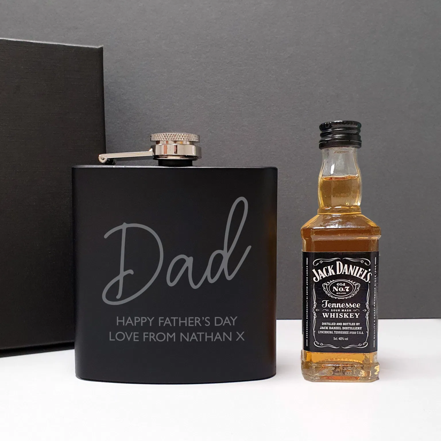Personalised Hip Flask Gift Set. A sleek matte black 6oz hip flask engraved with the name "Dad" in script and the message "HAPPY FATHER’S DAY LOVE FROM NATHAN X," standing next to a 5cl miniature bottle of Jack Daniel's whisky and a black presentation gift box.