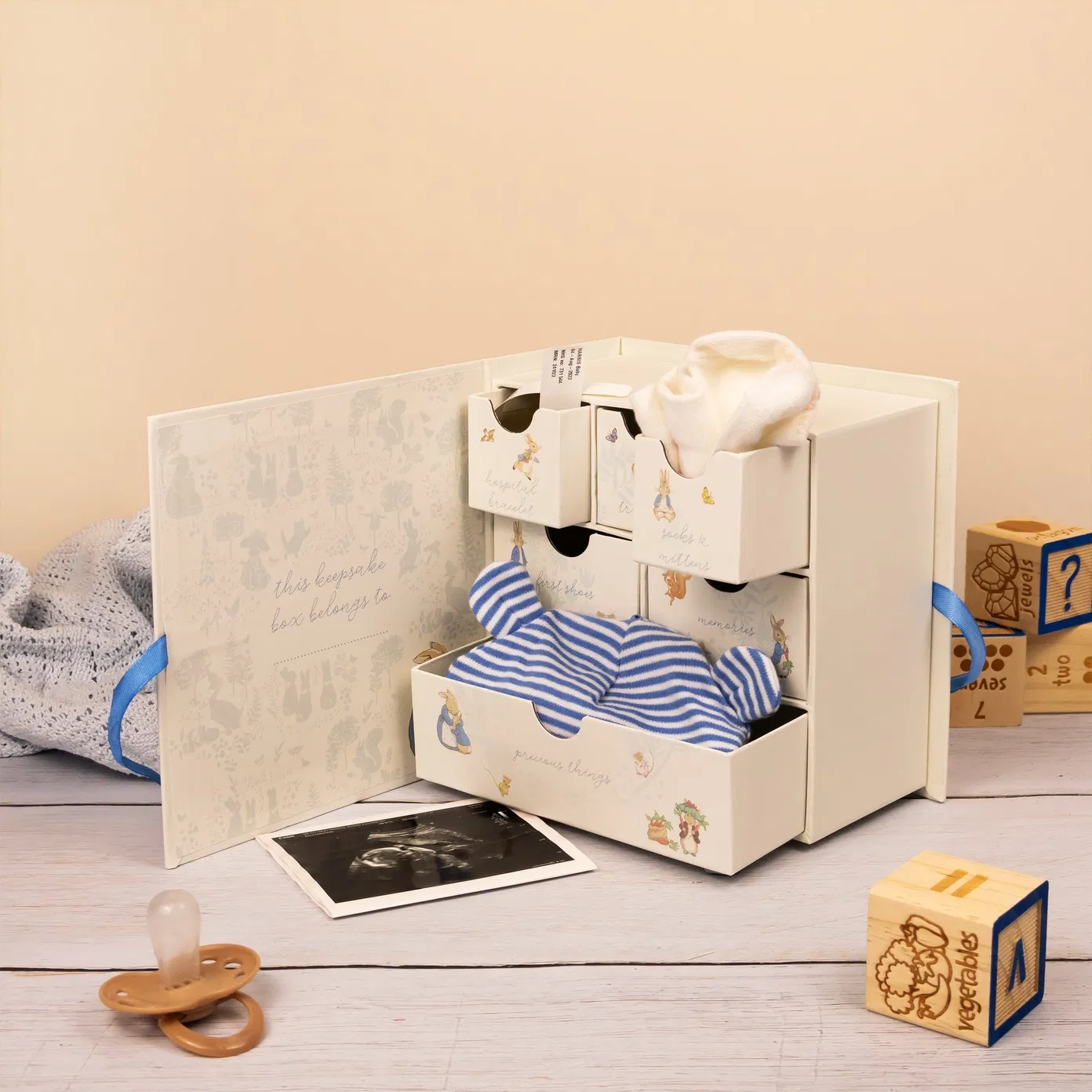 Open Peter Rabbit baby memento chest featuring six drawers labeled for hospital bracelets, socks, and first shoes. The box is decorated with classic Beatrix Potter illustrations, tied with a blue ribbon, and shown holding a baby hat, socks, and an ultrasound photo.