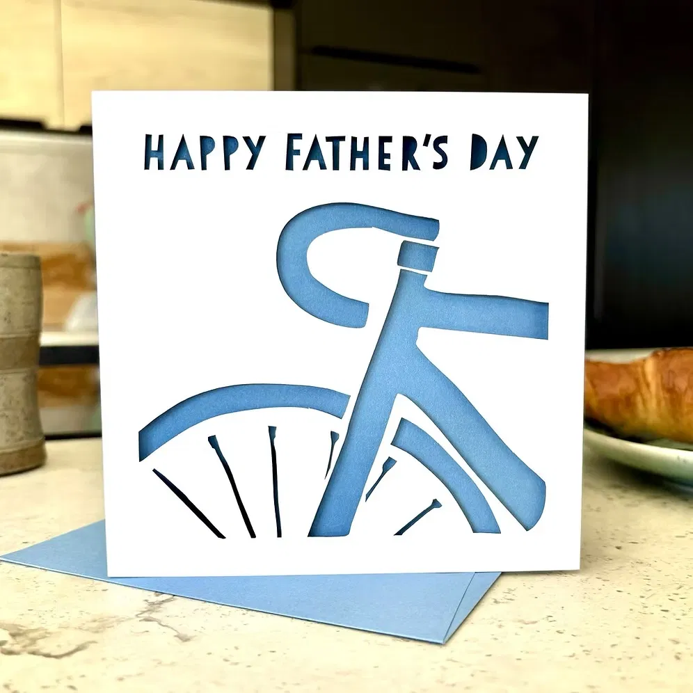 Cycling Father's Day Card: A square Father’s Day card featuring a white laser-cut silhouette of the front half of a road bicycle against a light blue background. The card reads "HAPPY FATHER'S DAY" in stencil-style lettering and is held over a matching blue envelope next to a coffee cup and croissant.