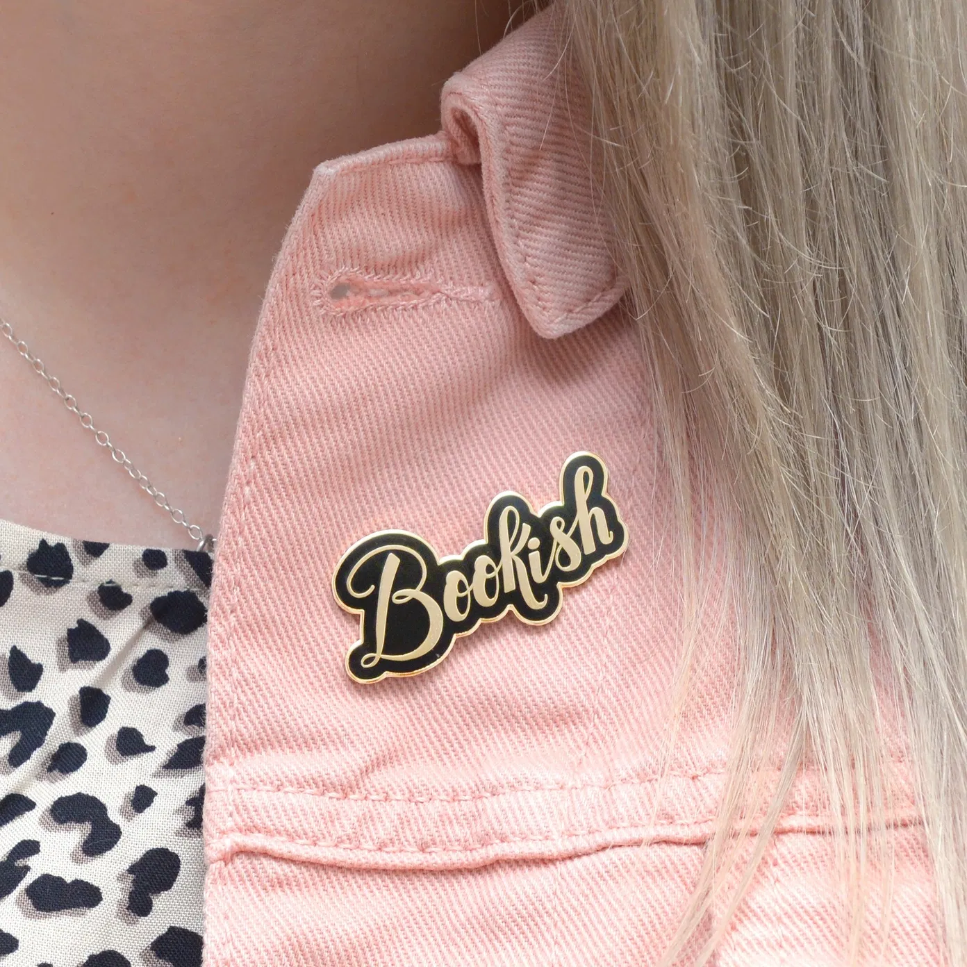 A close-up photograph of a person wearing a pink denim jacket, featuring a shiny black and gold hard enamel lapel pin that reads "Bookish" in elegant cursive script.