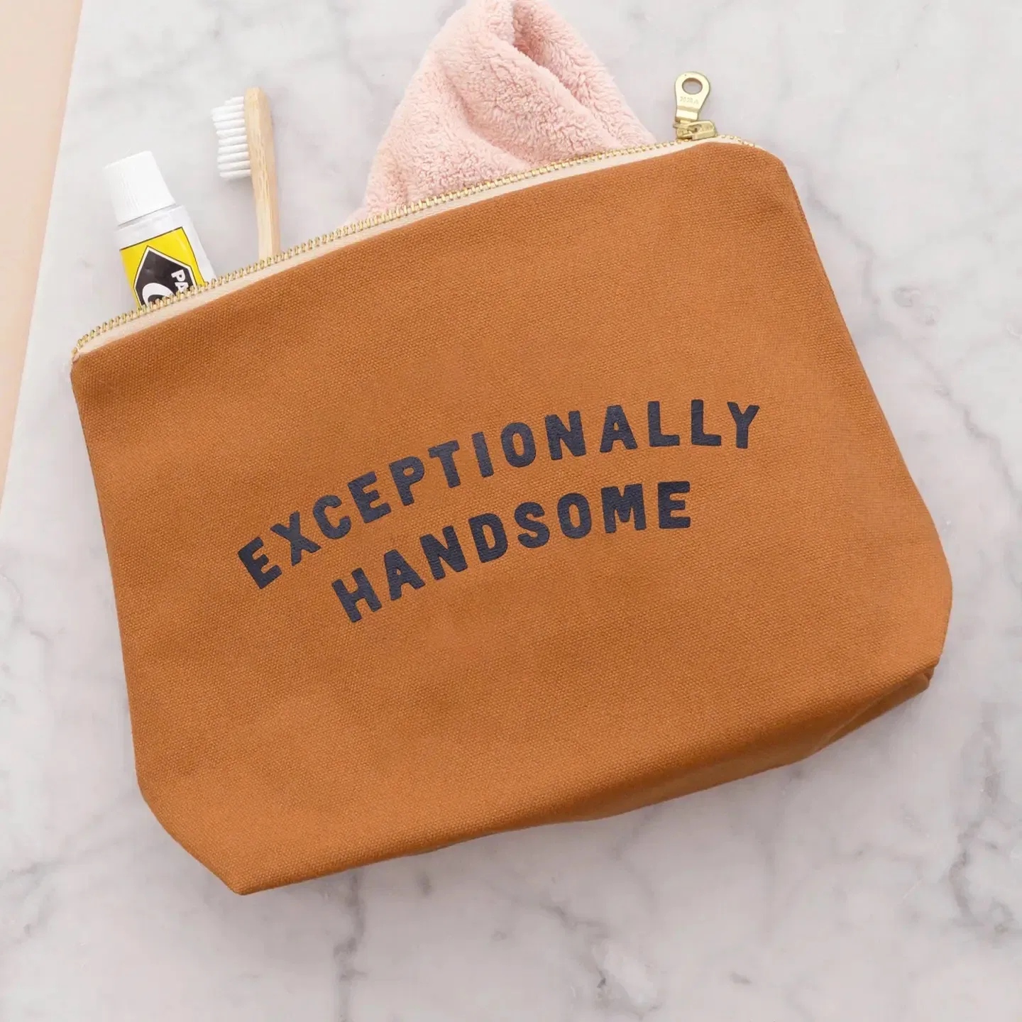A flat-lay shot of a tan canvas wash bag with 'Exceptionally Handsome' printed in navy blue, sitting on a marble surface with a toothbrush, toothpaste, and a pink face cloth peeking out from the gold zip opening.