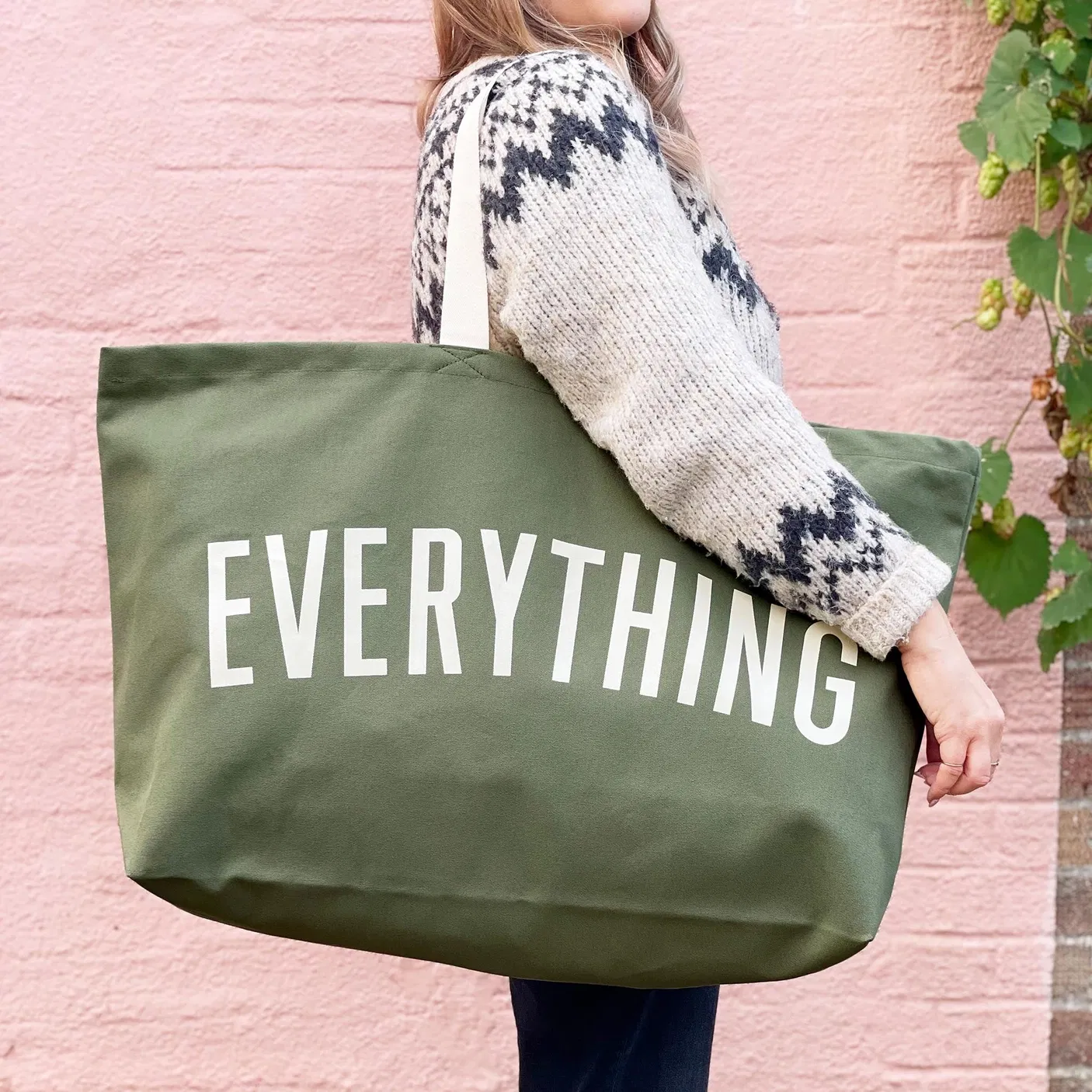 A person wearing a patterned sweater carrying an extra-large khaki green canvas tote bag that reads "EVERYTHING" in bold off-white lettering, set against a pink wall background.