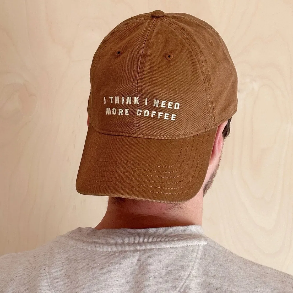 A back-facing view of a man wearing a tan brown cotton baseball cap. The text "I THINK I NEED MORE COFFEE" is embroidered in off-white on the back of the cap above the strap.