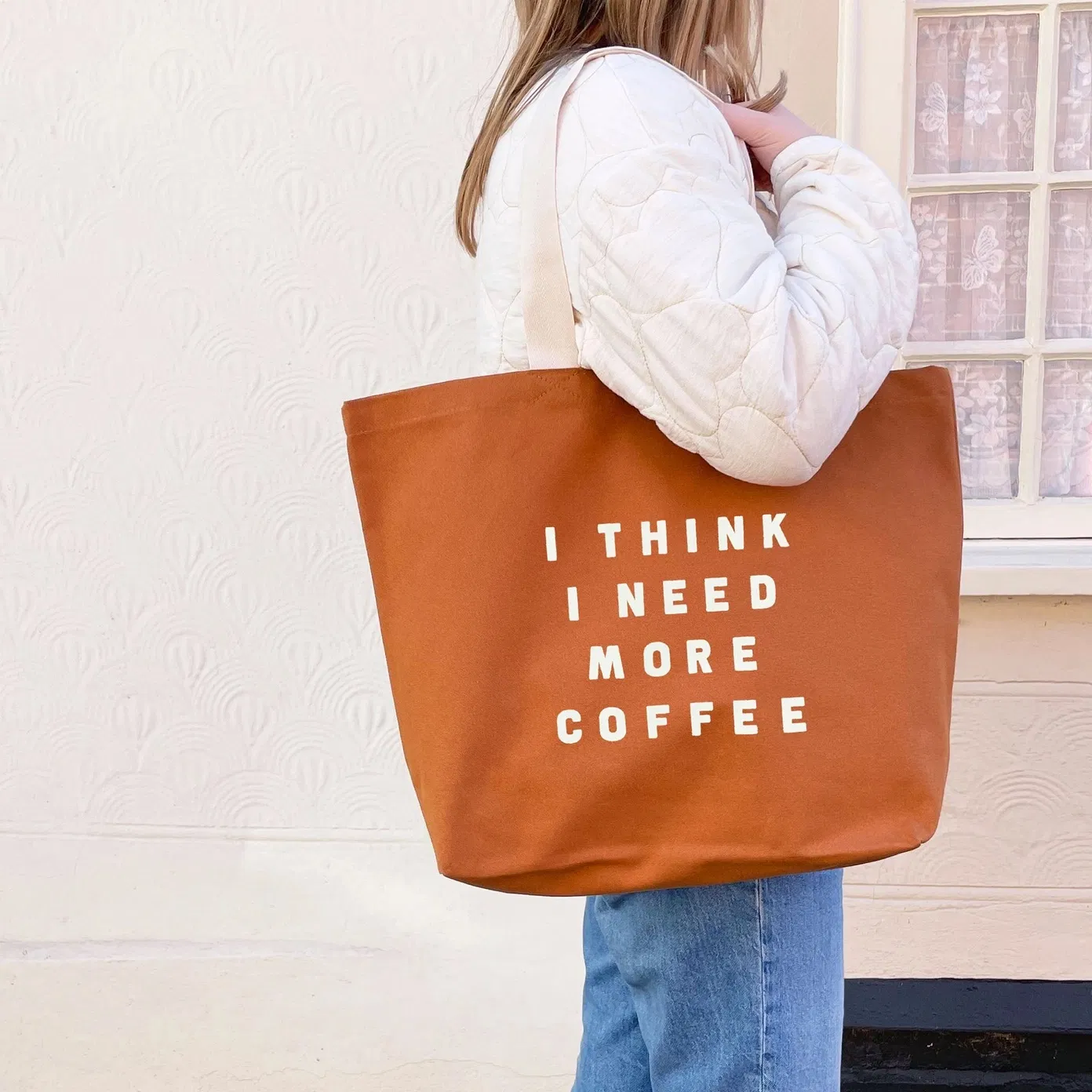 A person wearing a white quilted jacket carries a tan, 100% cotton canvas tote bag featuring the text 'I THINK I NEED MORE COFFEE' in off-white lettering.