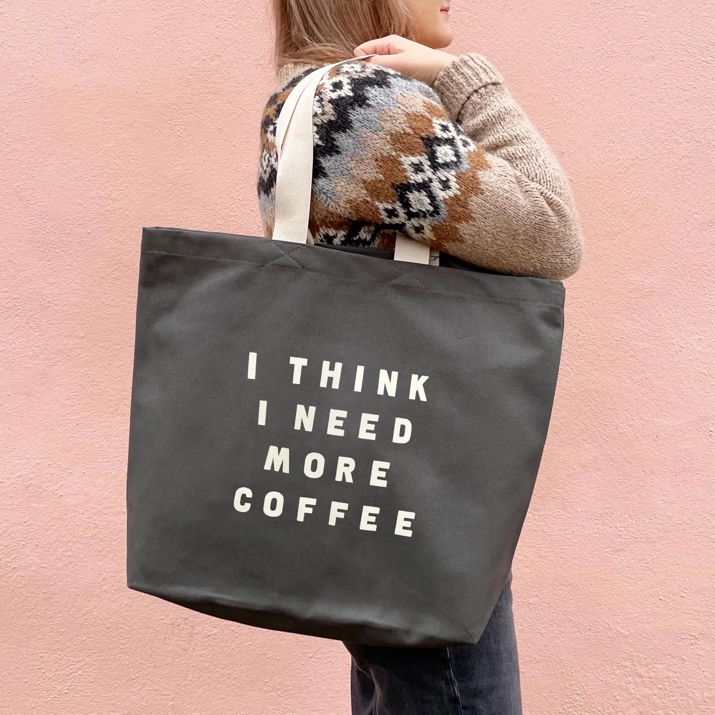 A person wearing a patterned knit sweater carries a dark grey, 100% cotton canvas tote bag featuring the text 'I THINK I NEED MORE COFFEE' in off-white lettering.