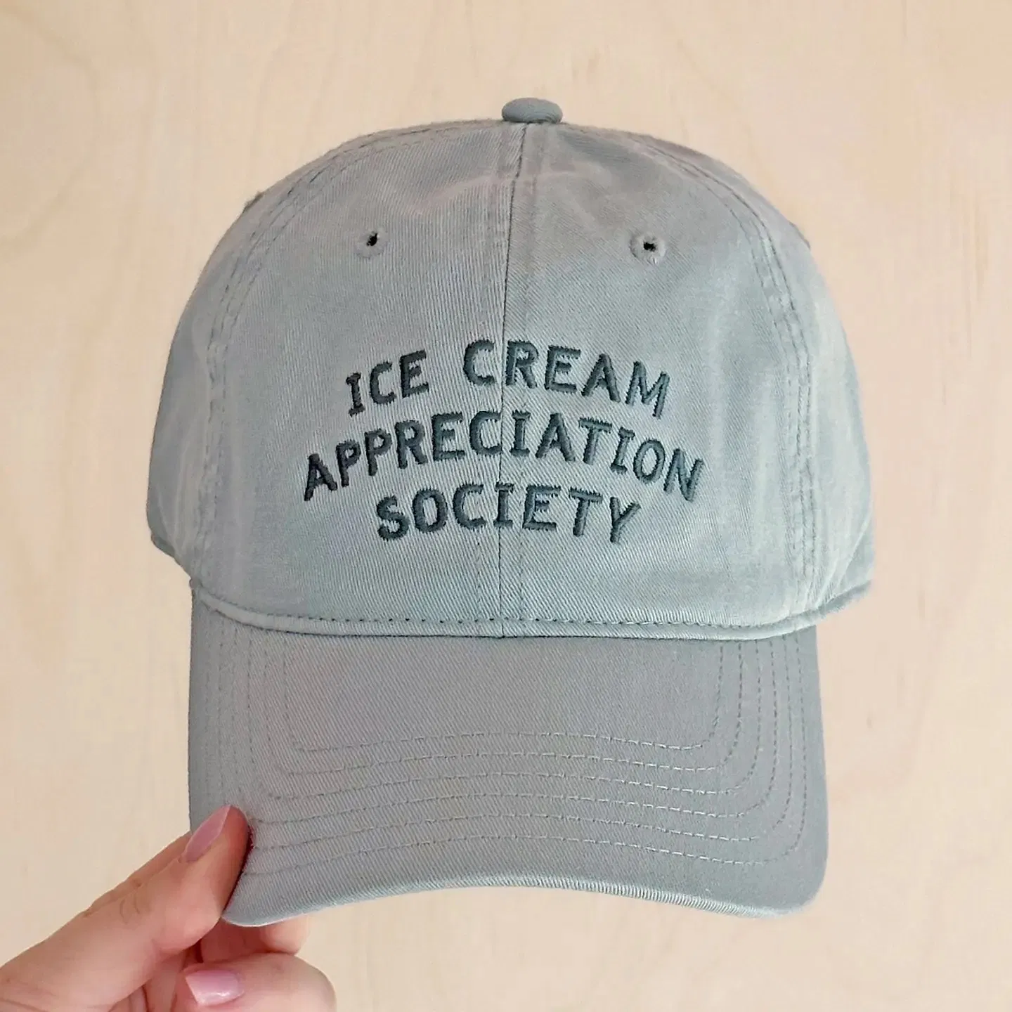 Hand holding a pale vintage blue cotton baseball cap with Ice Cream Appreciation Society embroidered on the front, set against a light wooden background.