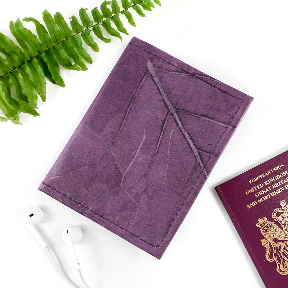 A bold purple Leaf Leather Passport Cover showcasing organic vein patterns, laid on a clean white surface next to a green fern leaf and a pair of white earphones.