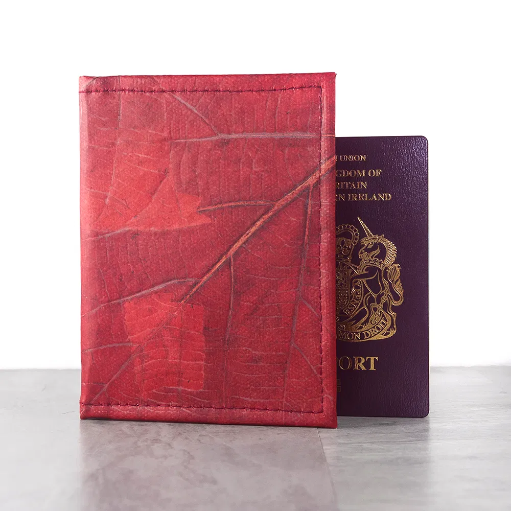 A red Leaf Leather Passport Cover made from dried, natural leaves, standing upright next to a UK passport on a light wooden surface.