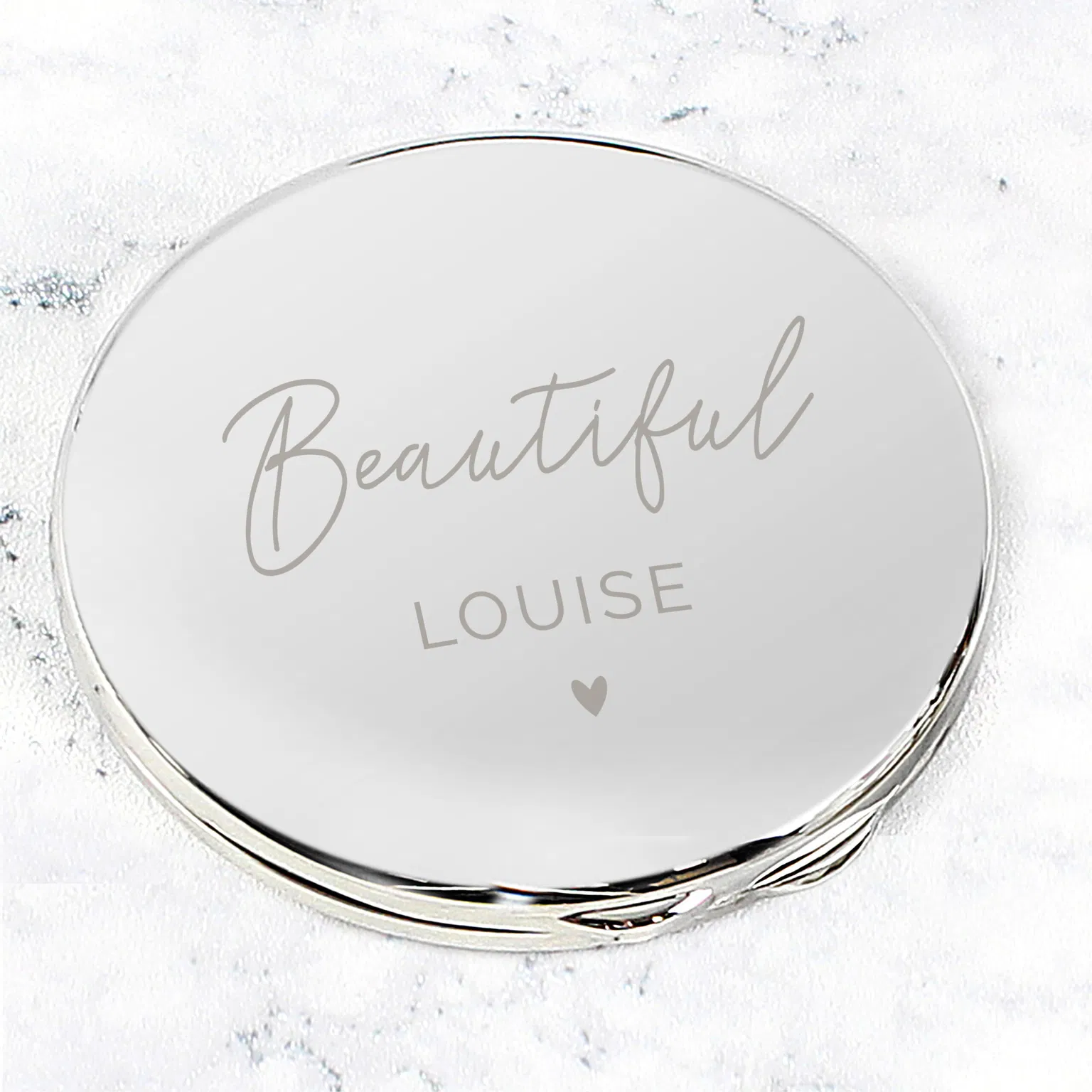 A studio-lit, high-angle close-up of a round, silver compact mirror resting on a white marble background. The mirror is engraved with the word "Beautiful" in elegant script, followed by the name "LOUISE" and a small heart icon.