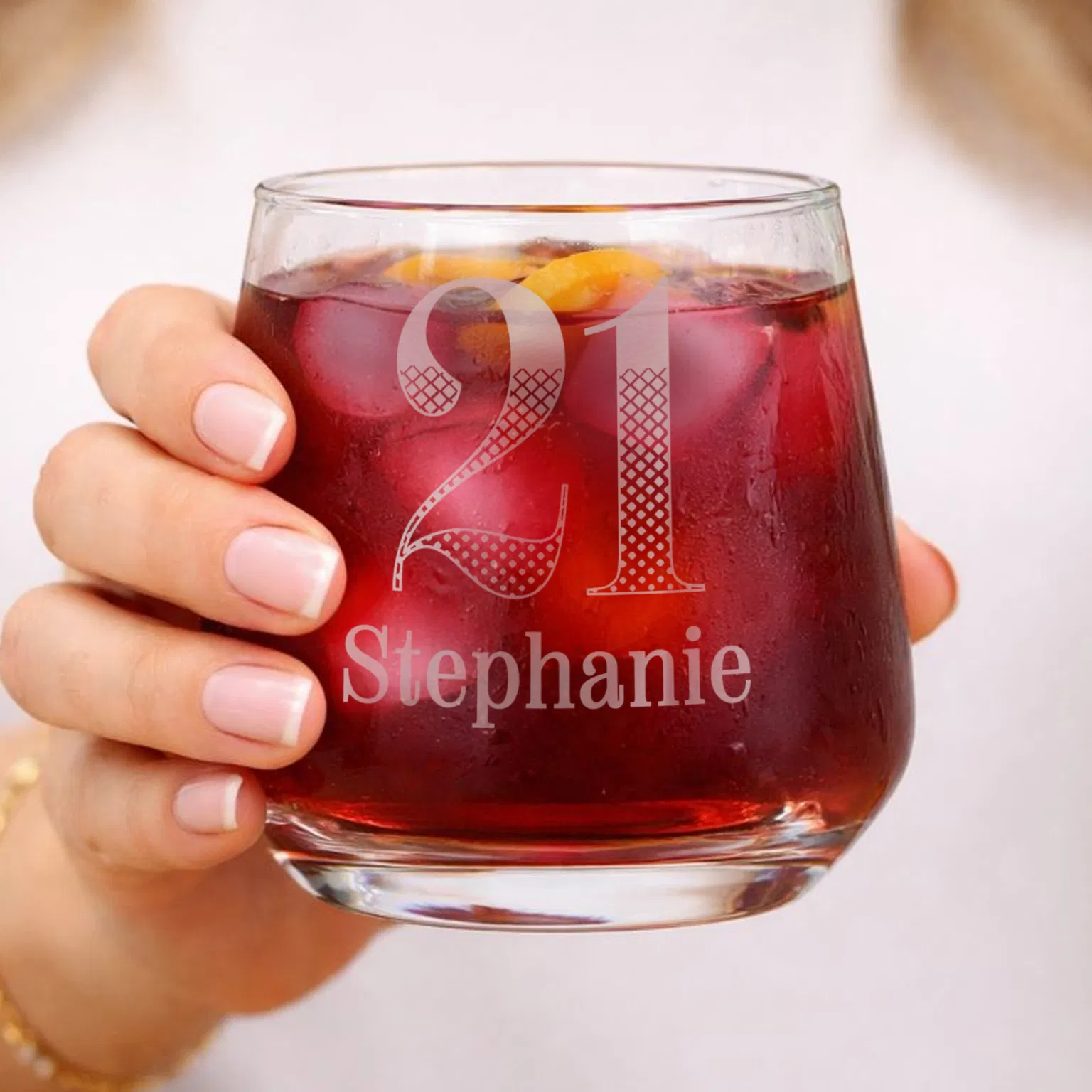 Personalised 21st Birthday Tumbler: A hand holding a custom-engraved glass tumbler filled with a dark red cocktail. The glass features a large, decorative patterned "21" and the name "Stephanie" engraved beneath it.
