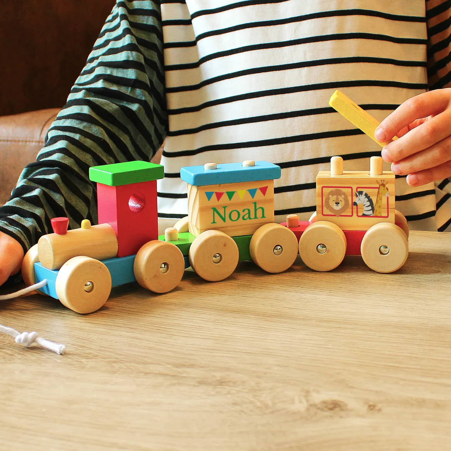A wooden toy train with a circus theme sitting on a wooden table. A child's hand is holding a block above the train, which is personalised with the name "Noah".