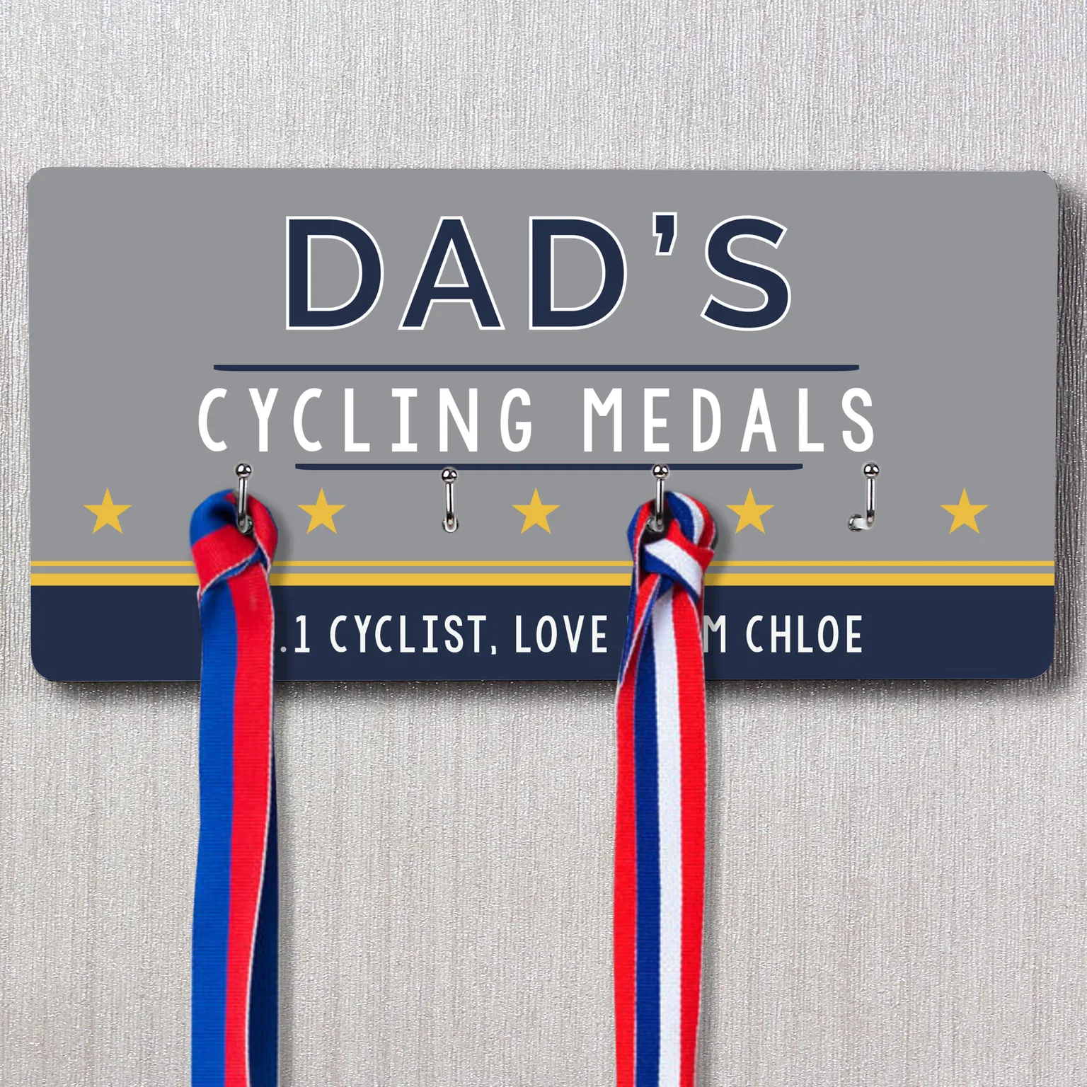 Personalised Dad's Achievement Medal Hooks: A grey and navy rectangular personalised medal rack hanging on a wall, featuring the text "DAD'S CYCLING MEDALS," five yellow stars, and a personalised message below reading "OUR #1 CYCLIST, LOVE CHLOE." Two medals with red, white, and blue ribbons are shown hanging from the hooks.