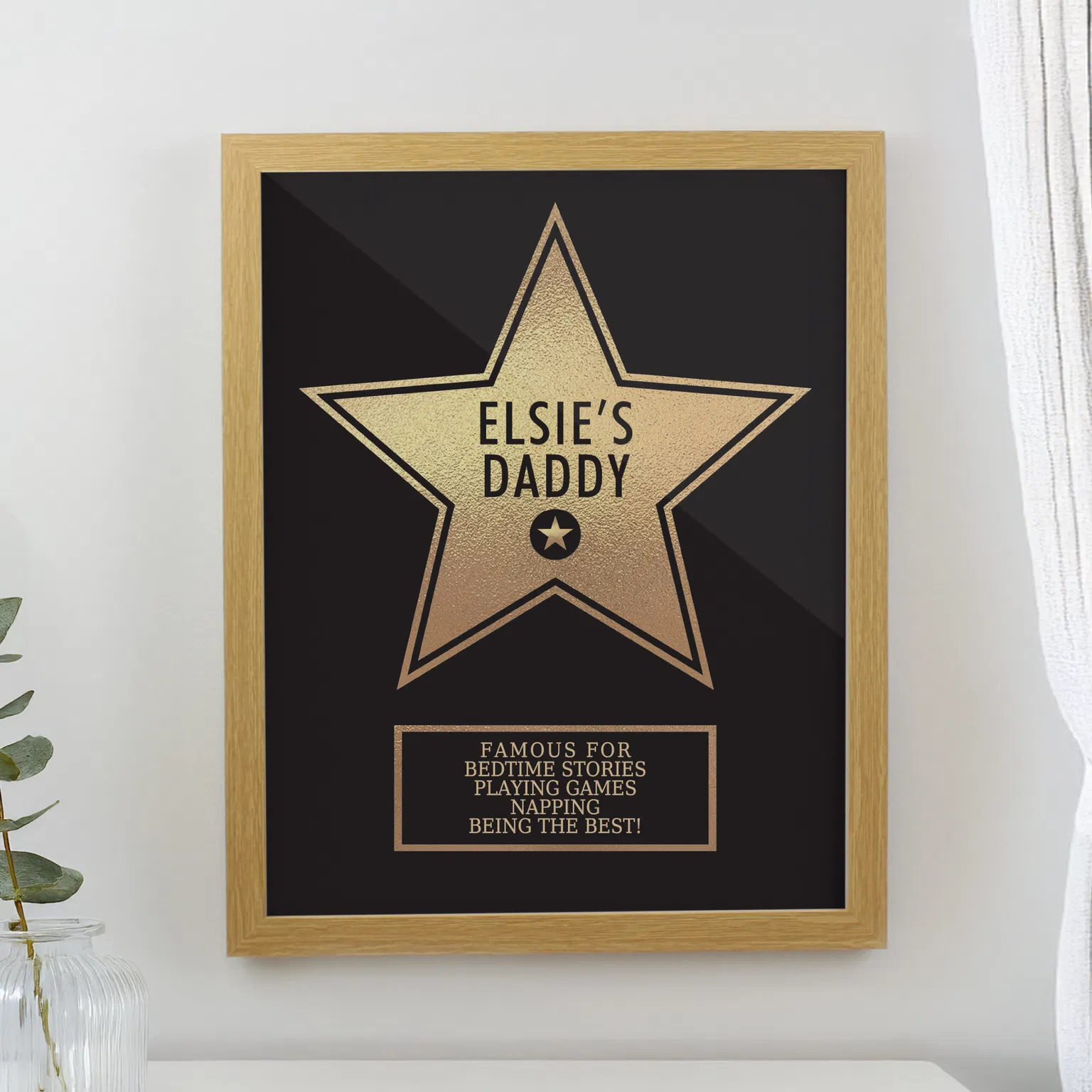 Personalised Father's Day Star Award Frame: A gold star award design in a light wood frame, hanging on a white wall. The star features the text "Elsie's Daddy" and a plaque listing things he is famous for, like "Bedtime Stories" and "Napping."