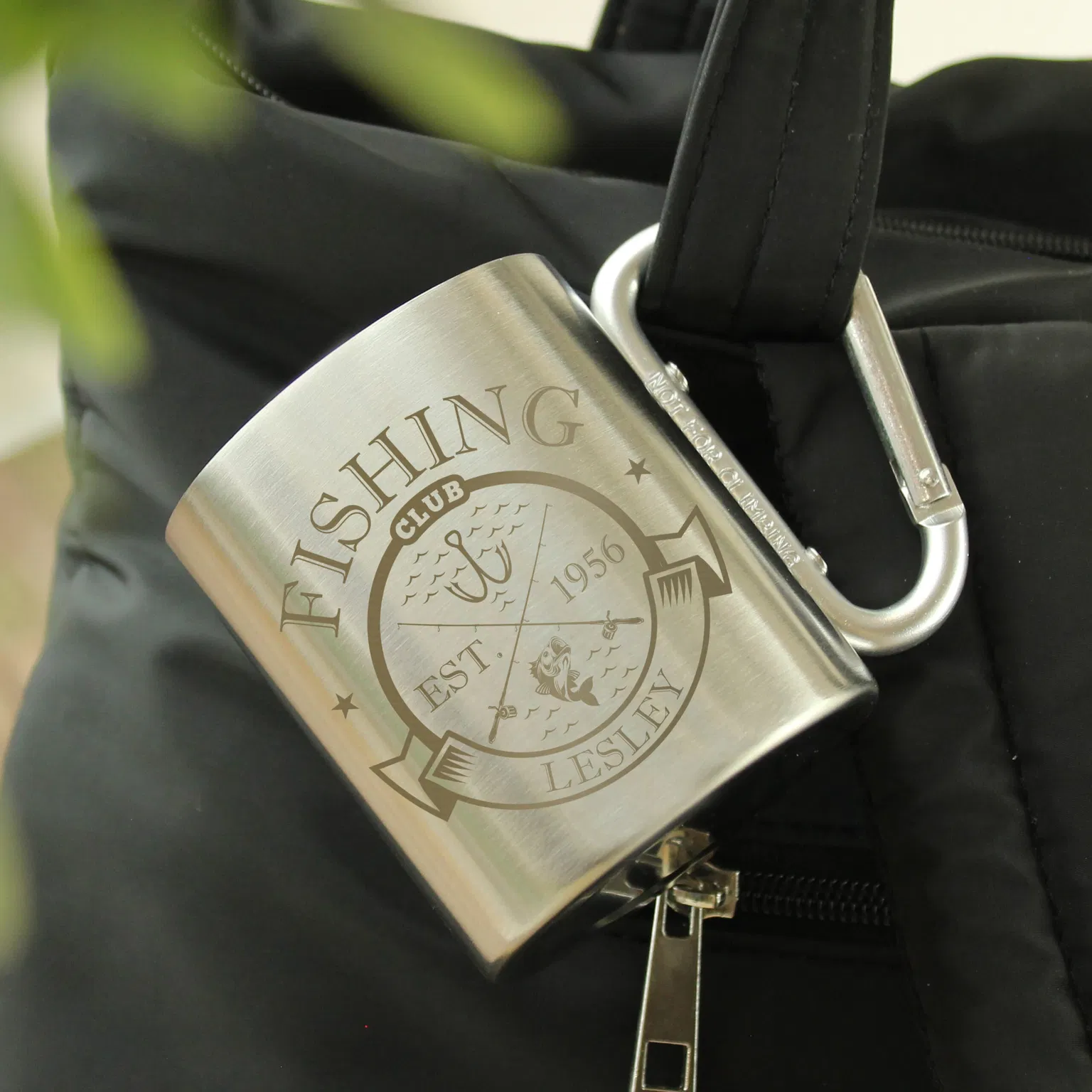 Personalised Fishing Club Mug: A silver stainless steel mug with a carabiner handle clipped to a black bag. The mug is engraved with a "Fishing Club" crest featuring a fish and hooks, personalised with the name "LESLEY" and the year "1956."