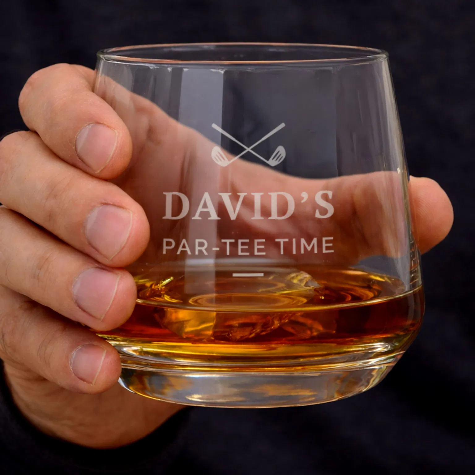 A hand holding a personalized glass whiskey tumbler filled with amber liquid and ice, featuring an engraved golf club logo and text that reads "David's Par-Tee Time."