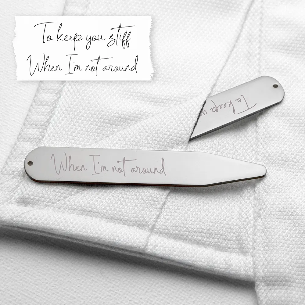 A close-up photograph of a pair of silver rhodium-plated collar stiffeners resting on a crisp white dress shirt. One stiffener is inserted into the collar fold, revealing the engraved handwriting text "When I'm not around." Above the shirt, a strip of white paper displays the original handwritten note: "To keep you stiff / When I'm not around".