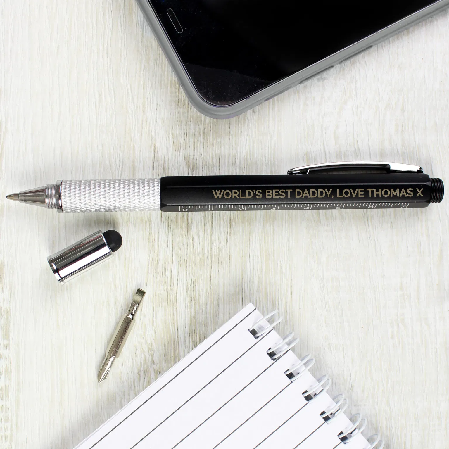 Personalised Multi Tool Pen: A black 7-in-1 multi-tool pen with an engraved personalised message in silver capital letters, showing the built-in spirit level, ruler markings, and screwdriver attachment on a wooden desk.