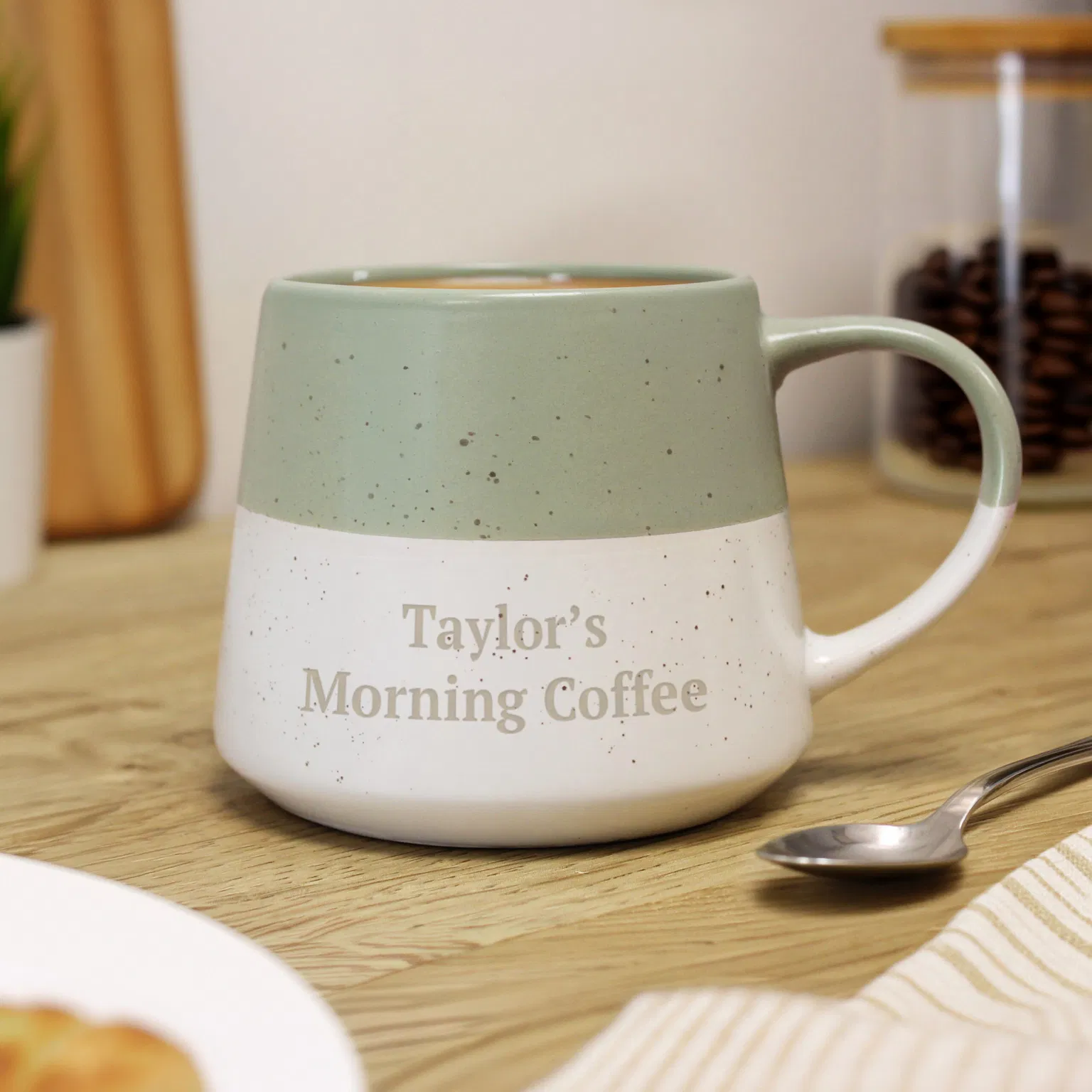 A two-tone, sage green and white speckled ceramic mug resting on a wooden surface, engraved with the text "Taylor's Morning Coffee".
