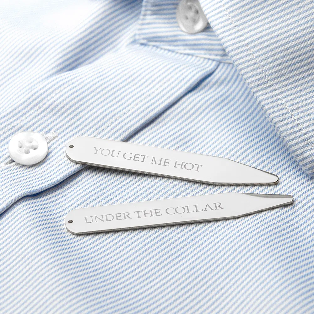A close-up studio shot of two silver rhodium-plated collar stiffeners resting on a crisp, light blue and white pinstriped men's dress shirt. The stiffener at the top is engraved with the text "YOU GET ME HOT" and the one below it reads "UNDER THE COLLAR". The shirt has a white button visible in the background.