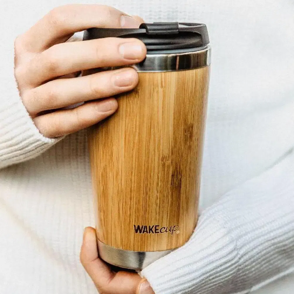 A person wearing a white knit jumper holding a WAKEcup reusable coffee cup made of natural bamboo with a black leak-proof lid and stainless steel trim.