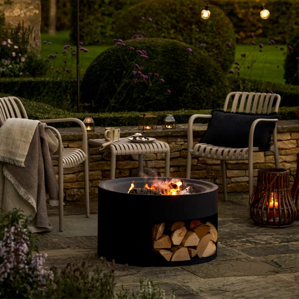 Matte black round steel fire pit with a log store base and glowing fire on a stone patio with garden seating.