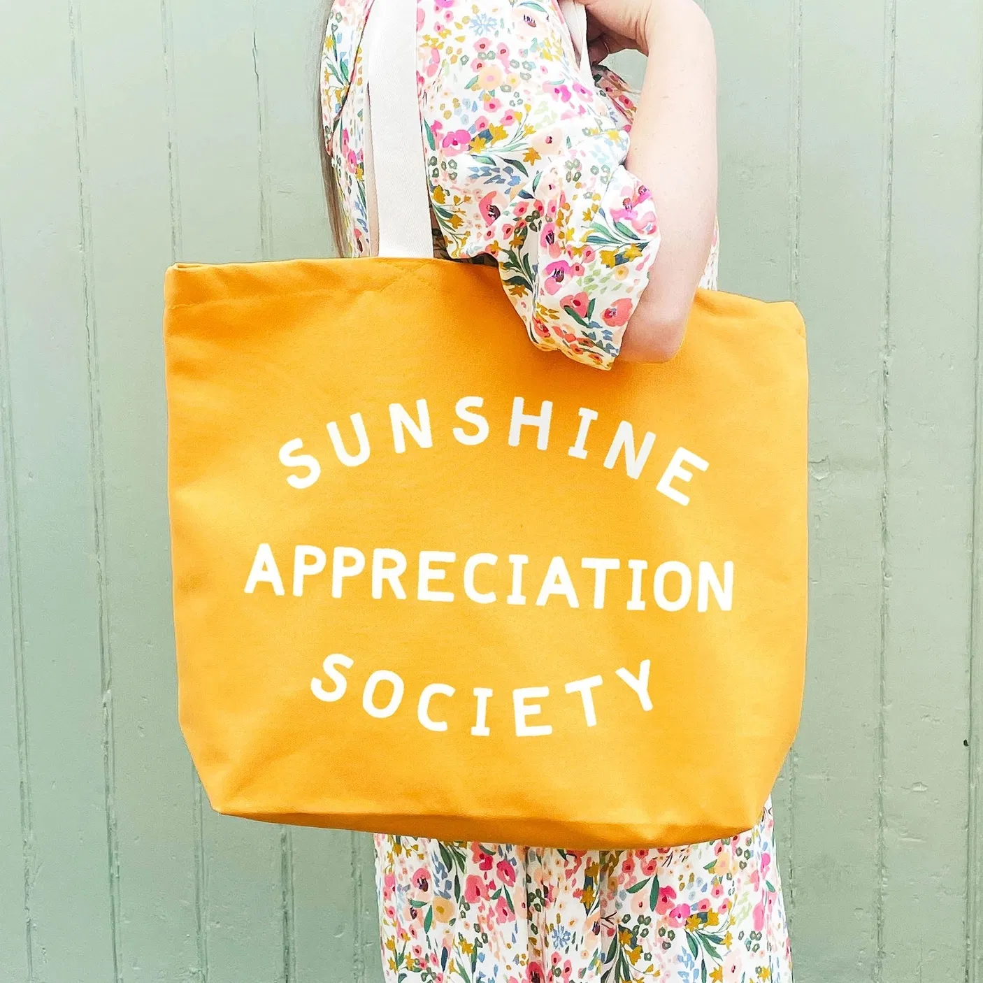 A person wearing a floral dress carrying a vibrant yellow canvas tote bag that reads "SUNSHINE APPRECIATION SOCIETY" in bold off-white lettering, against a matching green wooden background.