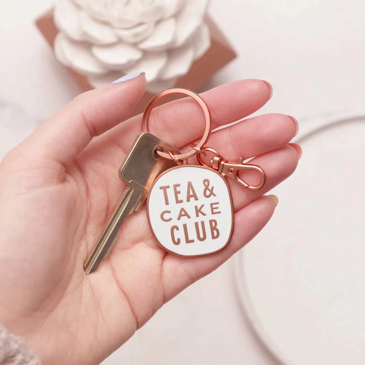 A close-up shot of a hand holding a round white enamel keyring with copper text that reads Tea and Cake Club, featuring a matching copper lobster clasp and keyring.