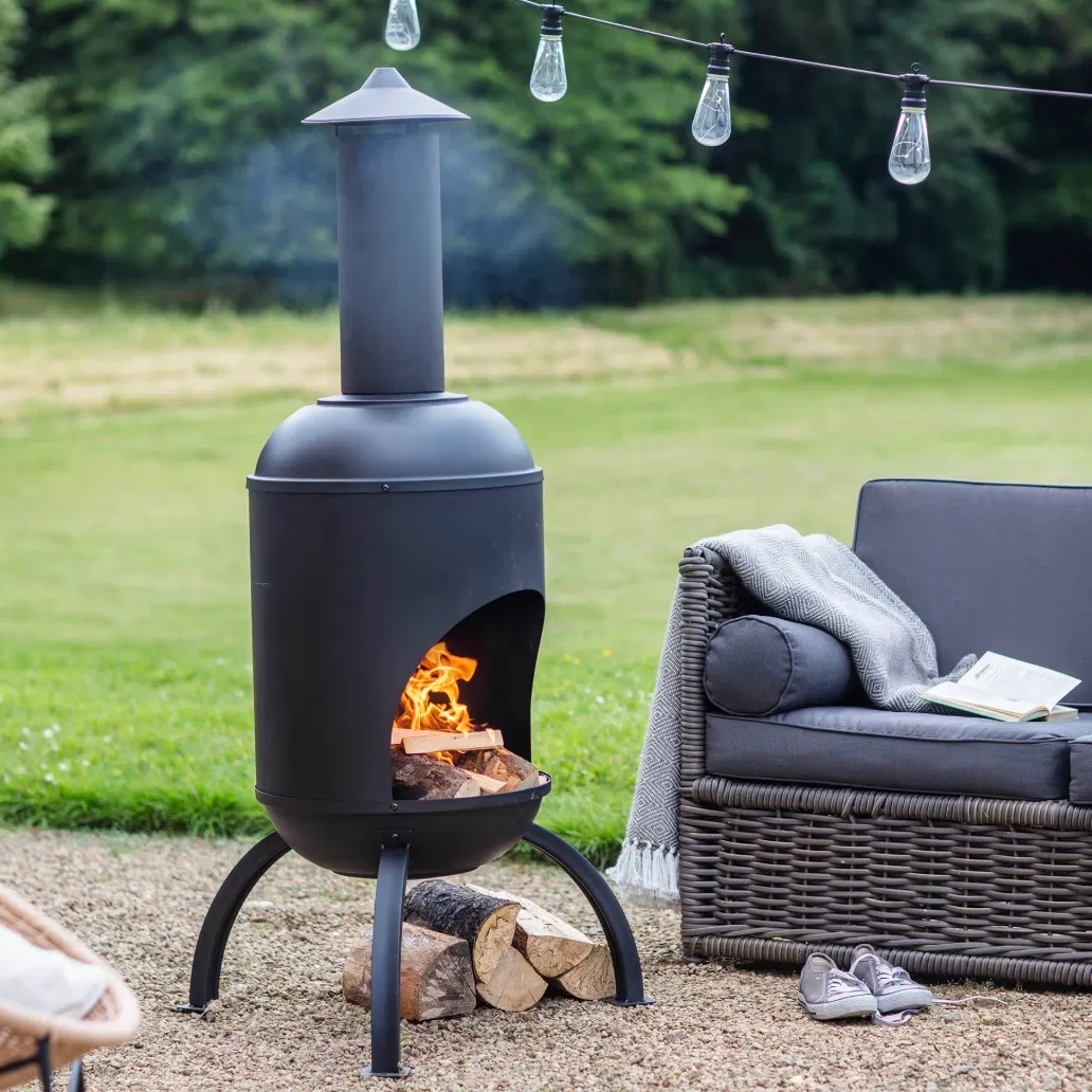 A Black Steel Chiminea with a lit wood fire, positioned on a gravel patio next to a modern grey garden sofa and outdoor string lights.