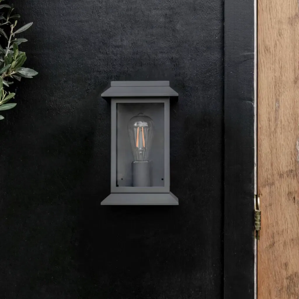 Modern carbon steel lantern wall light with a matte finish and glass framing, mounted on a dark exterior wall next to a natural wood door frame.