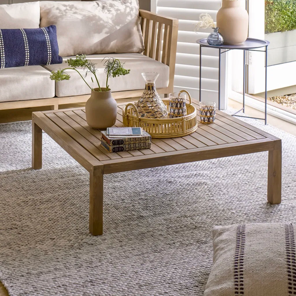 Square whitewash acacia wood coffee table styled with a tray and vase on a rug in a sunroom.