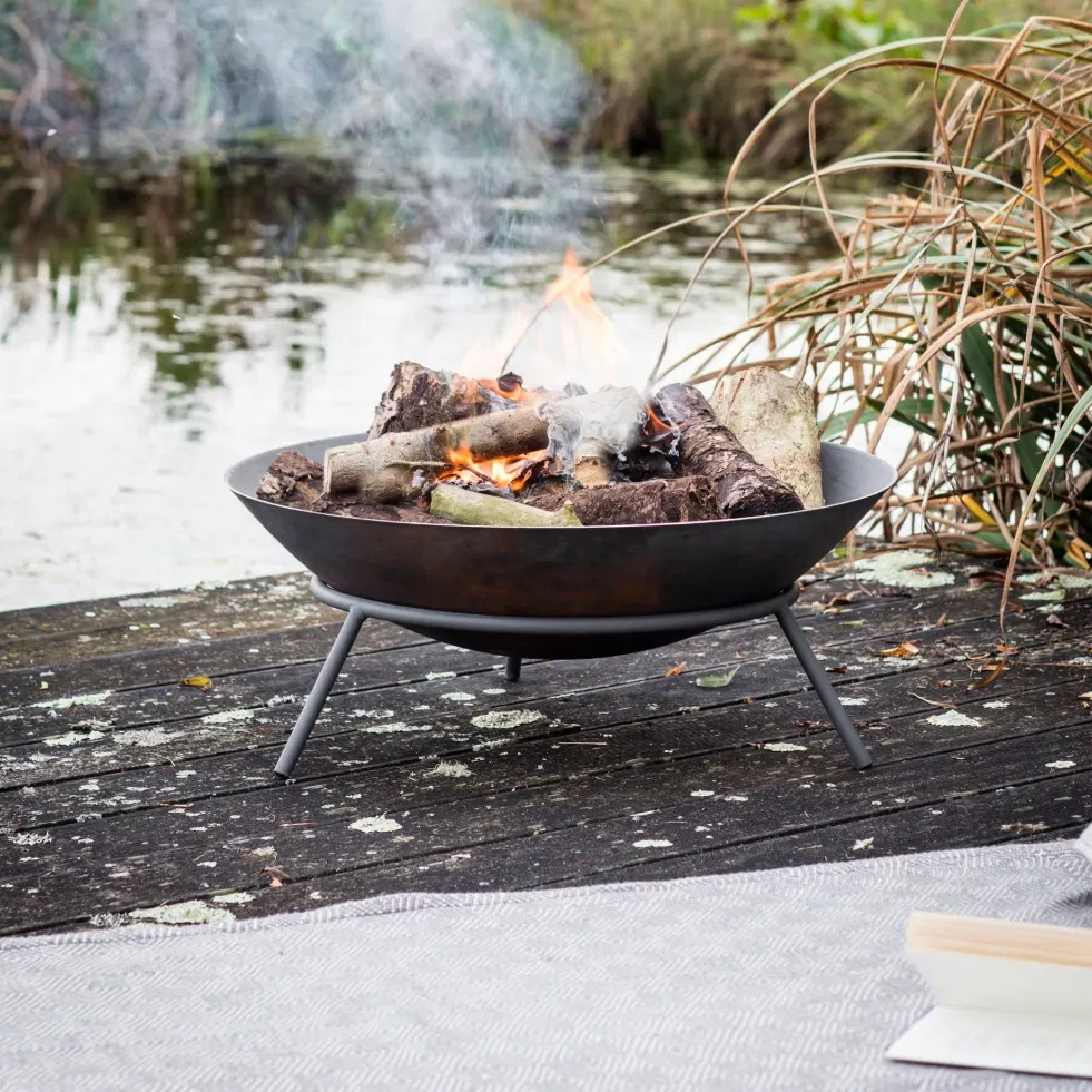 An Idbury Cast Iron Fire Pit with a log fire burning, positioned on a wooden jetty by a pond with a grey throw and book nearby.