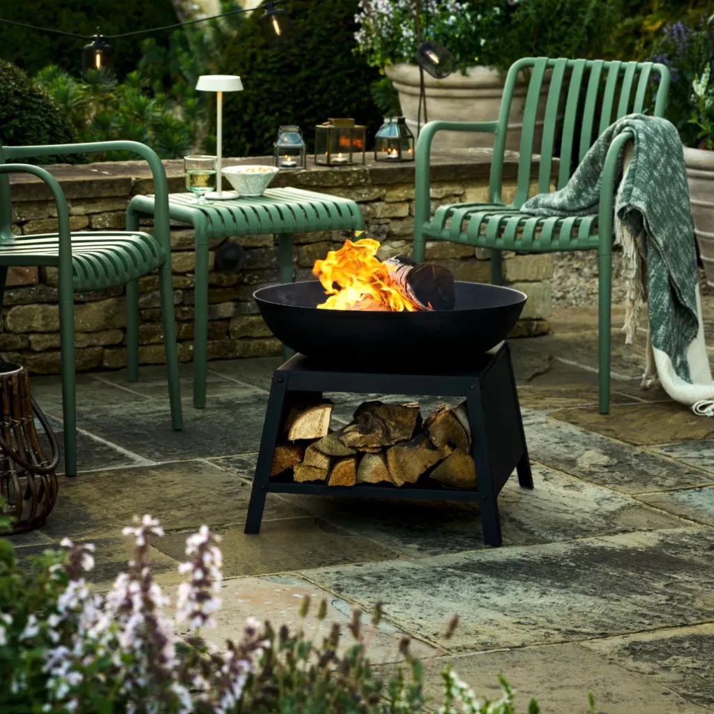 Matte black steel fire bowl with a burning log, featuring an integrated base for log storage, styled on a stone patio between two green garden chairs.