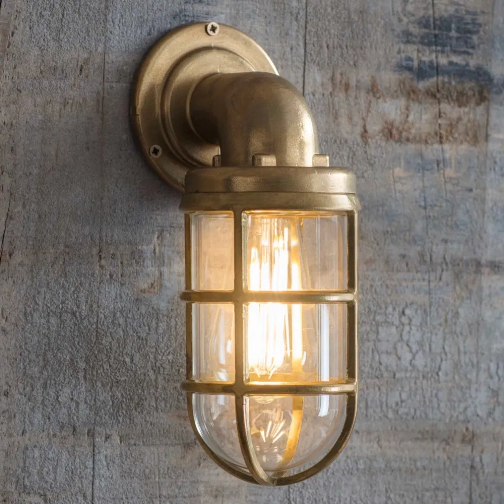 Nautical raw cast brass down light with an industrial cage design and lit filament bulb, mounted on a weathered wood wall.