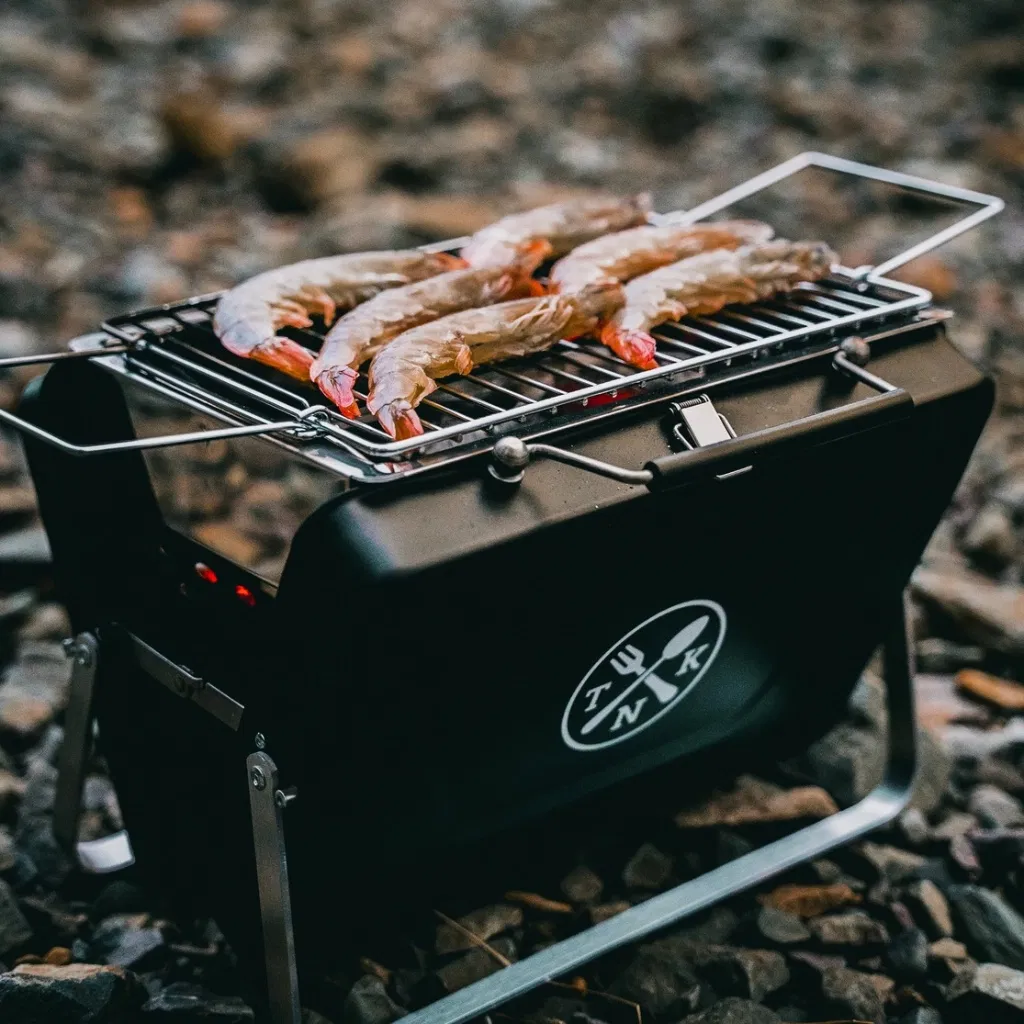 Portable Notebook BBQ with fresh king prawns grilling on a rocky beach.