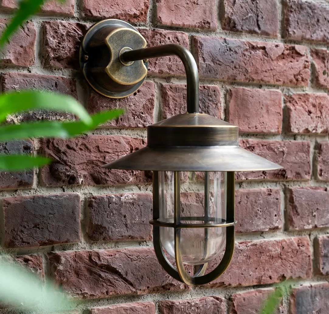 Antique brass outdoor wall light with a storm lamp-inspired cage and clear glass shade, mounted on a red brick wall.