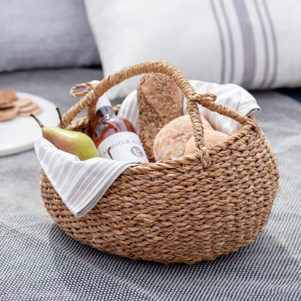 Natural seagrass picnic basket filled with fresh bread, wine, and fruit on a picnic rug.