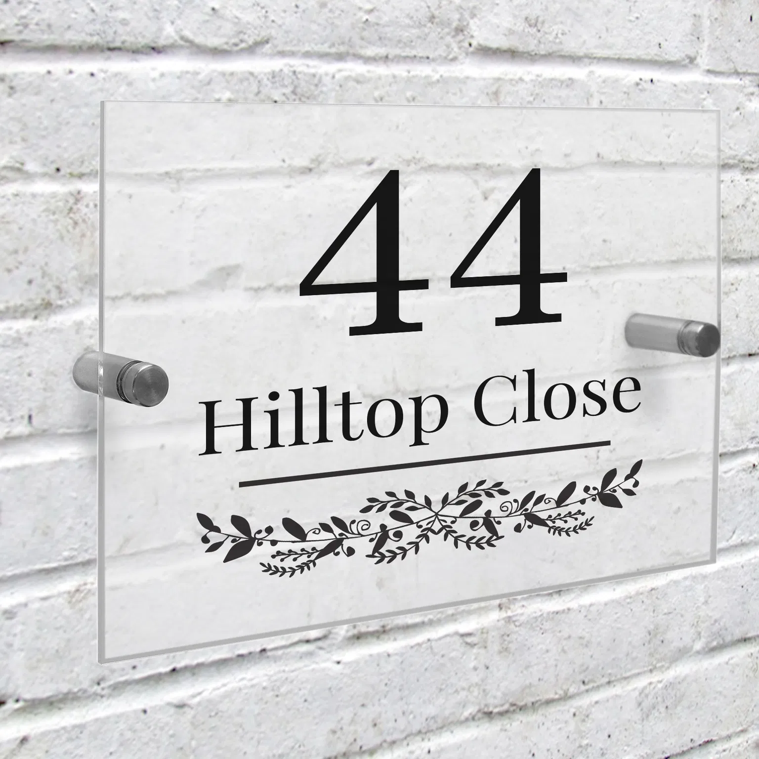 A clear rectangular acrylic house sign is mounted onto a white-painted brick wall using two silver metal stand-off fixings. The sign features a large black number '44' above the custom text 'Hilltop Close' and an elegant botanical leaf garland design.