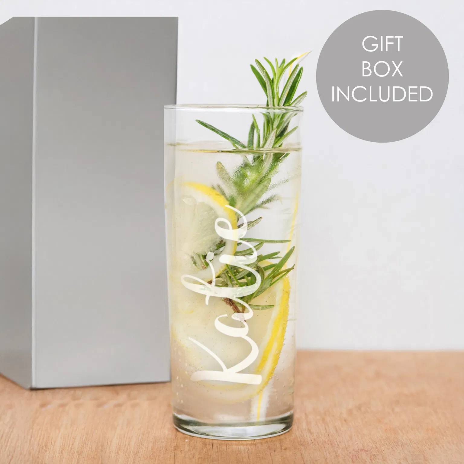 A clear glass hi ball tumbler filled with a clear carbonated drink, ice, lemon slices, and a fresh rosemary sprig sits on a wooden table. The name 'Katie' is engraved vertically on the front of the glass in a white script font. A grey folding gift box is visible in the background next to a circular 'Gift Box Included' graphic.
