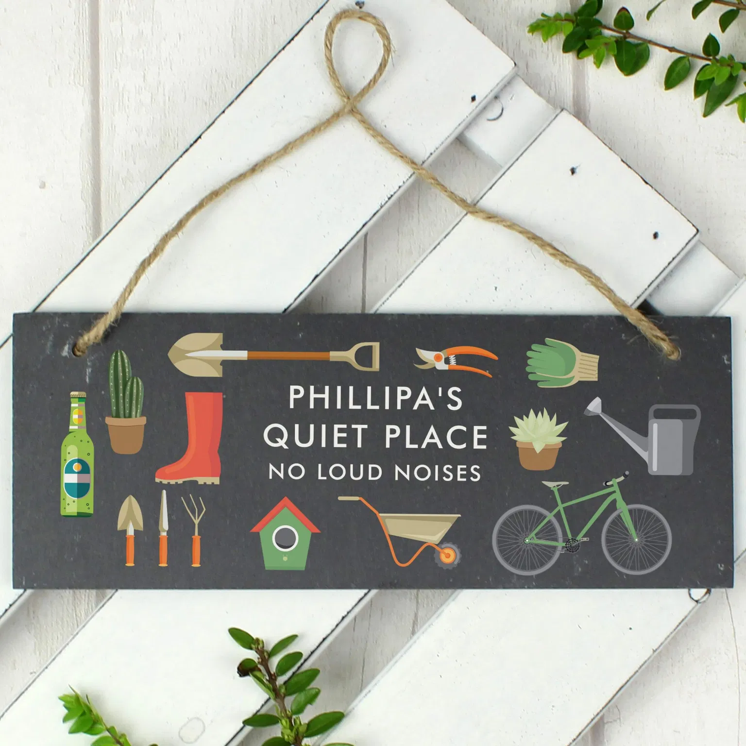 A rectangular hand-cut dark slate garden sign hangs flat against a white painted wooden slatted panel from a loop of rustic jute twine. The plaque is custom printed with fixed uppercase white text reading 'PHILLIPA'S QUIET PLACE, NO LOUD NOISES'. The wording is surrounded by colourful printed graphics of a watering can, wellington boot, bicycle, shovel, wheelbarrow, and small potted plants.