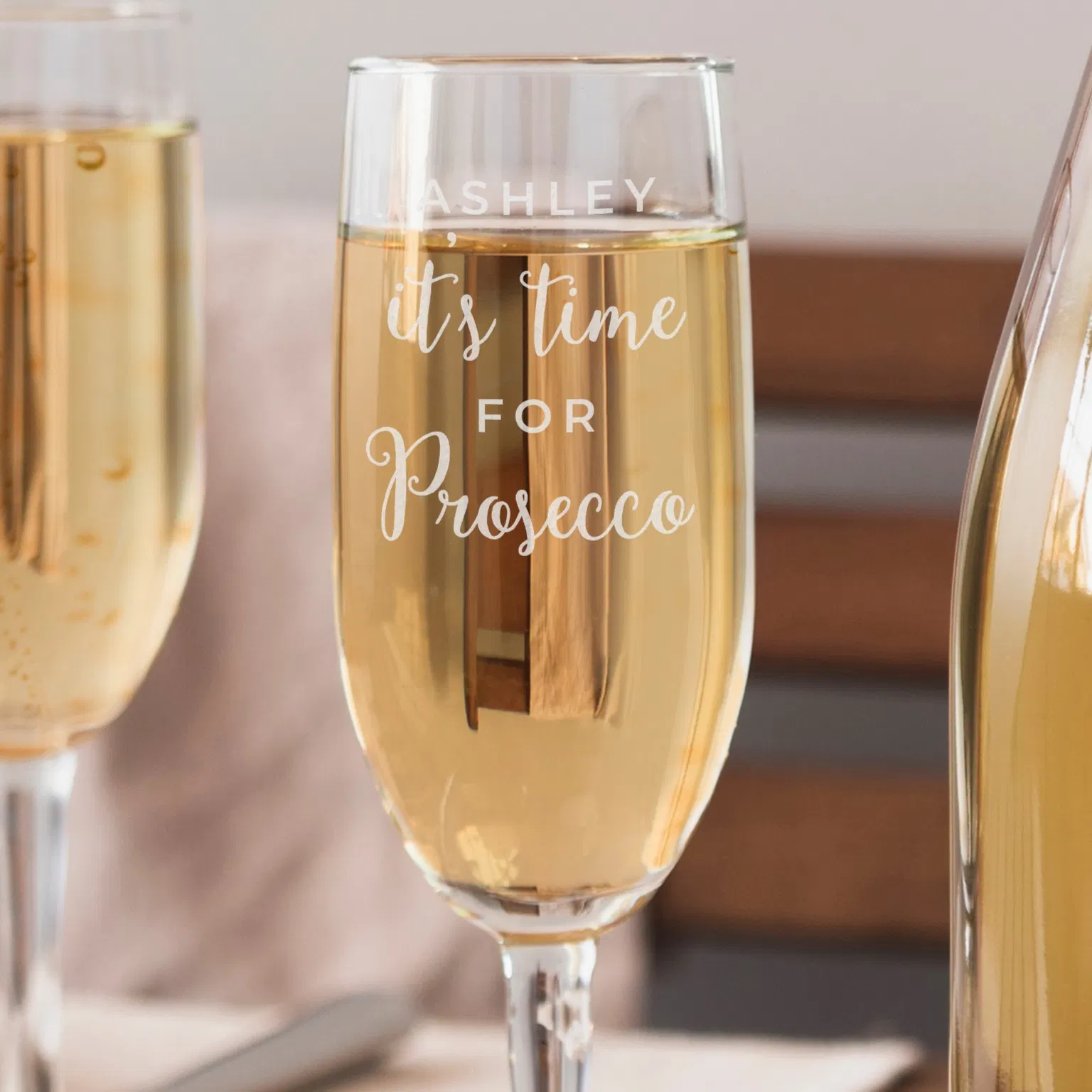 A clear glass champagne flute filled with bubbling prosecco sits on a table. The front of the glass is engraved with the name 'ASHLEY' in capital block letters, above the fixed script phrase 'it's time for Prosecco'. Another blurred flute and a prosecco bottle frame the background.