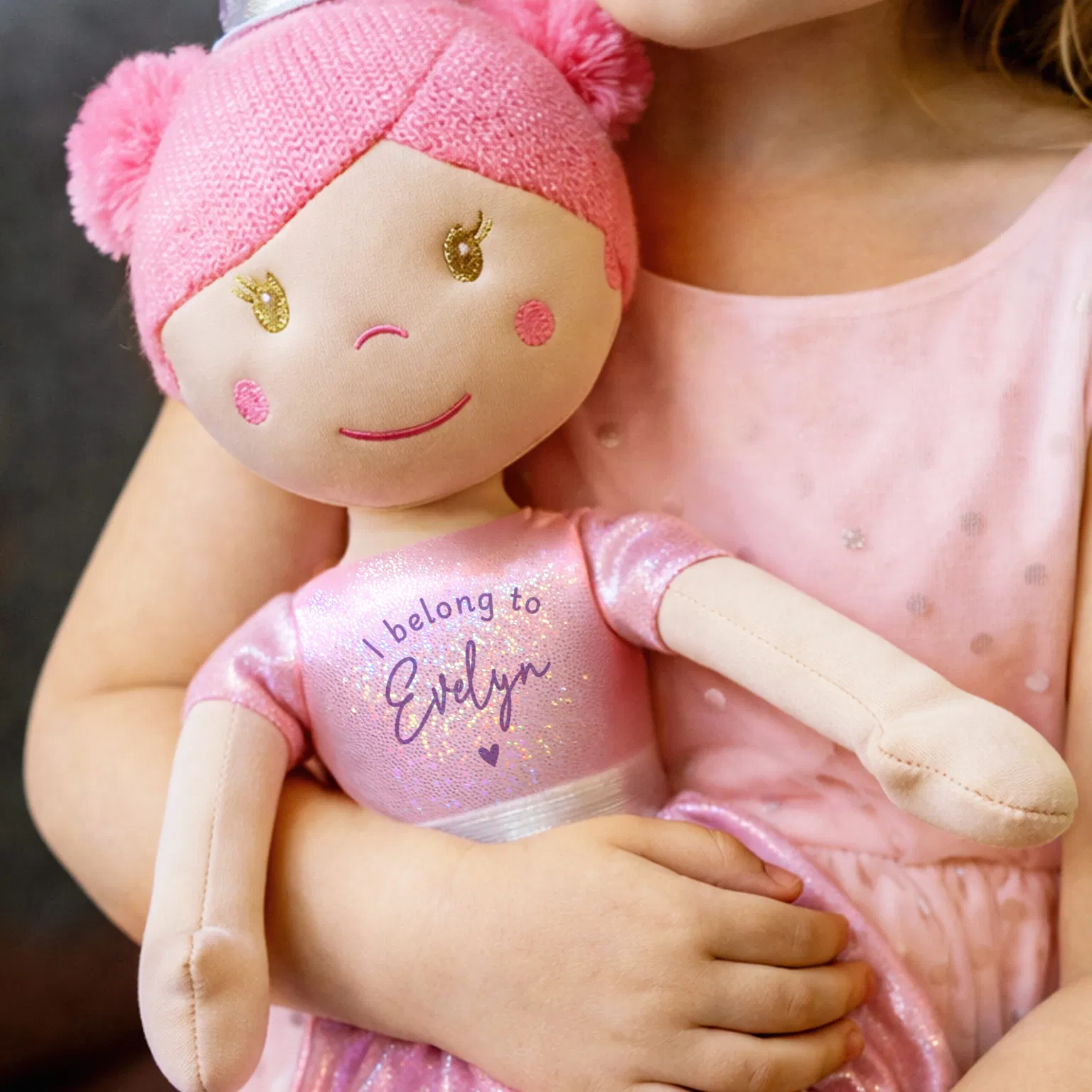 A close-up shot of a young girl in a pink sequined dress holding a plush princess rag doll. The doll has pink yarn hair, an embroidered smiling face, and wears a sparkling pink ballerina outfit text-printed with 'I belong to Evelyn' and a small purple heart.