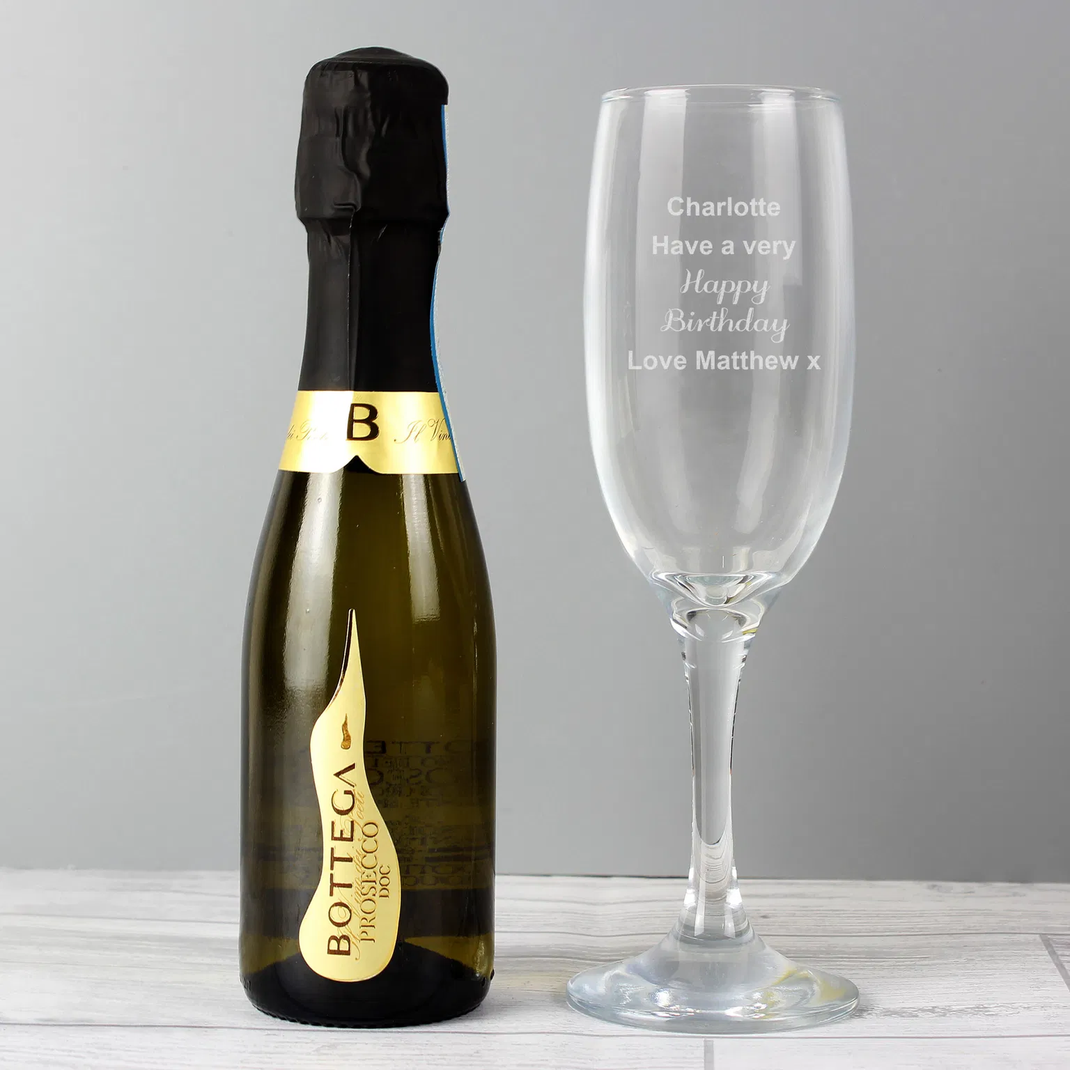 A clear glass champagne flute stands next to a miniature bottle of Bottega Prosecco DOC on a white wooden surface against a grey background. The glass bowl is laser-engraved with the case-sensitive text 'Charlotte, Have a very Happy Birthday, Love Matthew x'.