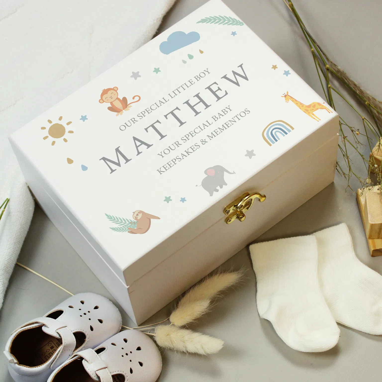 A rectangular hand-painted white wooden keepsake box with a gold metal clasp sits angled on a grey surface. The lid is printed with safari animals including an elephant, giraffe, monkey, and sloth around the capitalised custom text 'OUR SPECIAL LITTLE BOY, MATTHEW, YOUR SPECIAL BABY, KEEPSAKES & MEMENTOS'. White baby shoes and cream socks sit in the foreground.
