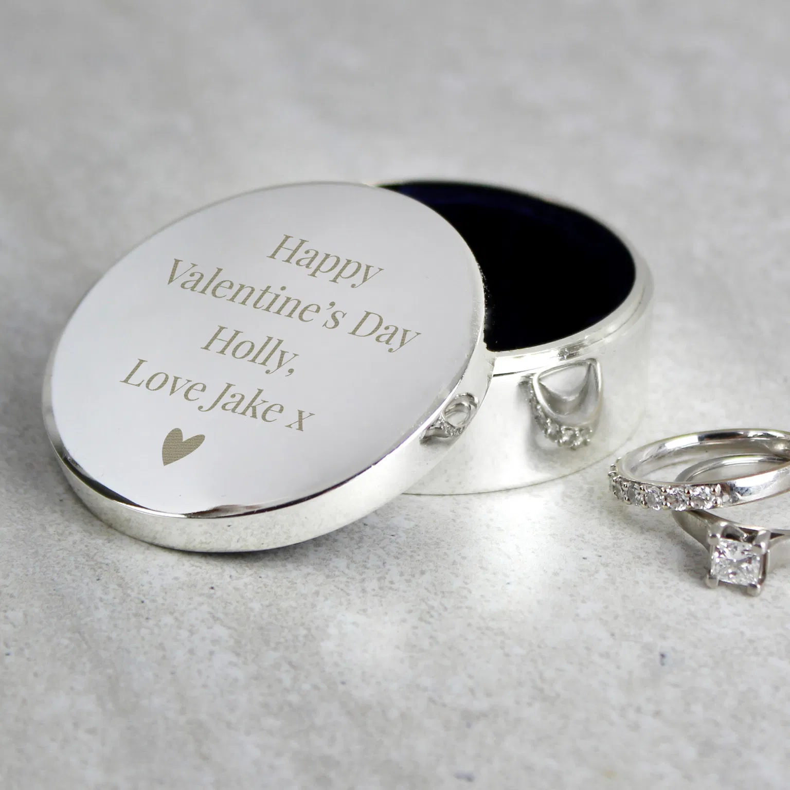 A round, mirror-polished silver ring box sits on a textured light grey surface with its lid propped open, revealing a dark velvet-lined interior. The lid is laser-engraved with 'Happy Valentine's Day, Holly, Love Jake x' above a small solid heart icon. A silver diamond engagement ring and matching wedding band sit beside it.