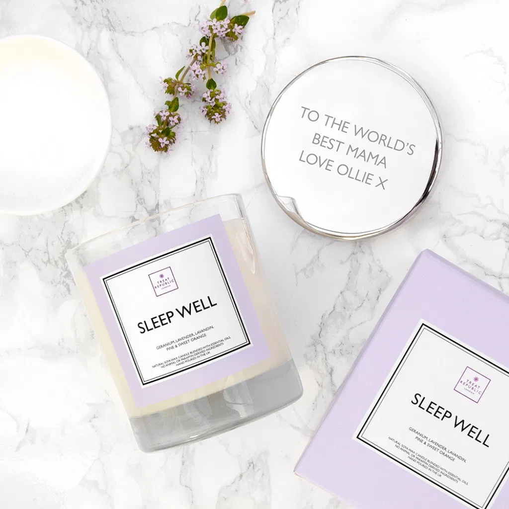 An overhead flat lay view of a clear glass 'Sleep Well' scented candle tumbler with a pastel purple label on a white marble surface. Beside it sits its polished silver metallic lid engraved with the text 'TO THE WORLD'S BEST MAMA LOVE OLLIE X'. A matching lilac gift box, a white ceramic dish, and a small sprig of purple flowers complete the styling.