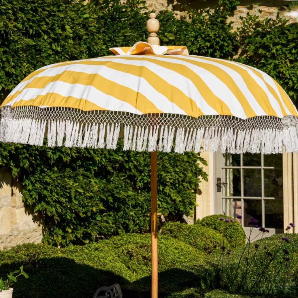 A luxury garden parasol with bright yellow and white canvas stripes and white cotton crochet fringe tassels. The umbrella has a central teak wood pole and stands on a manicured green lawn in front of a stone cottage wall draped in lush green ivy.