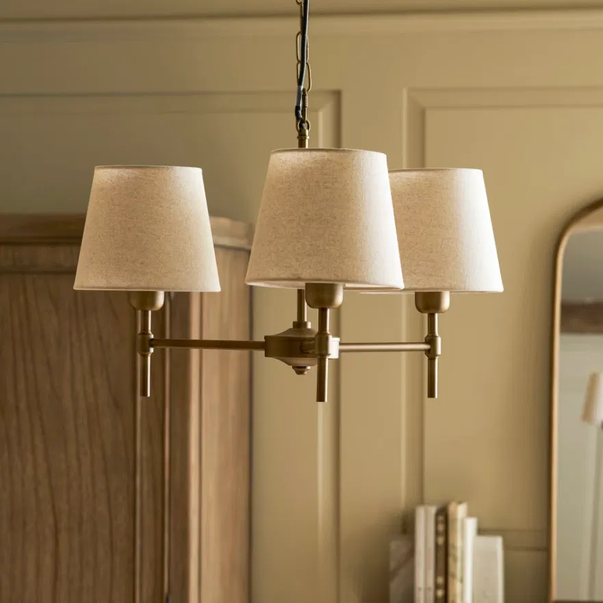 An antique bronze 3-light ceiling chandelier with cream linen shades styled in a room with an oak wardrobe and arched mirror by Abode Home & Gifts.